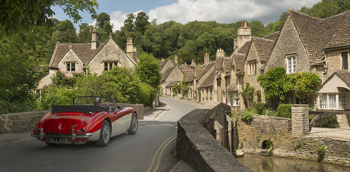 Red sports car drives over bridge outside cottages 